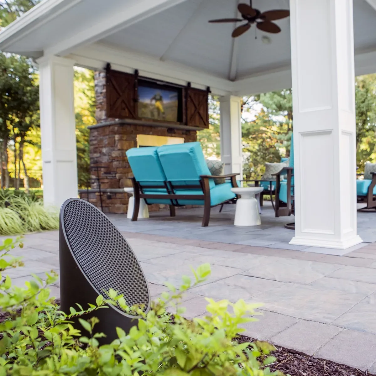 A Coastal Source bollard speaker in a planted bed with a covered pavilion and outdoor television behind