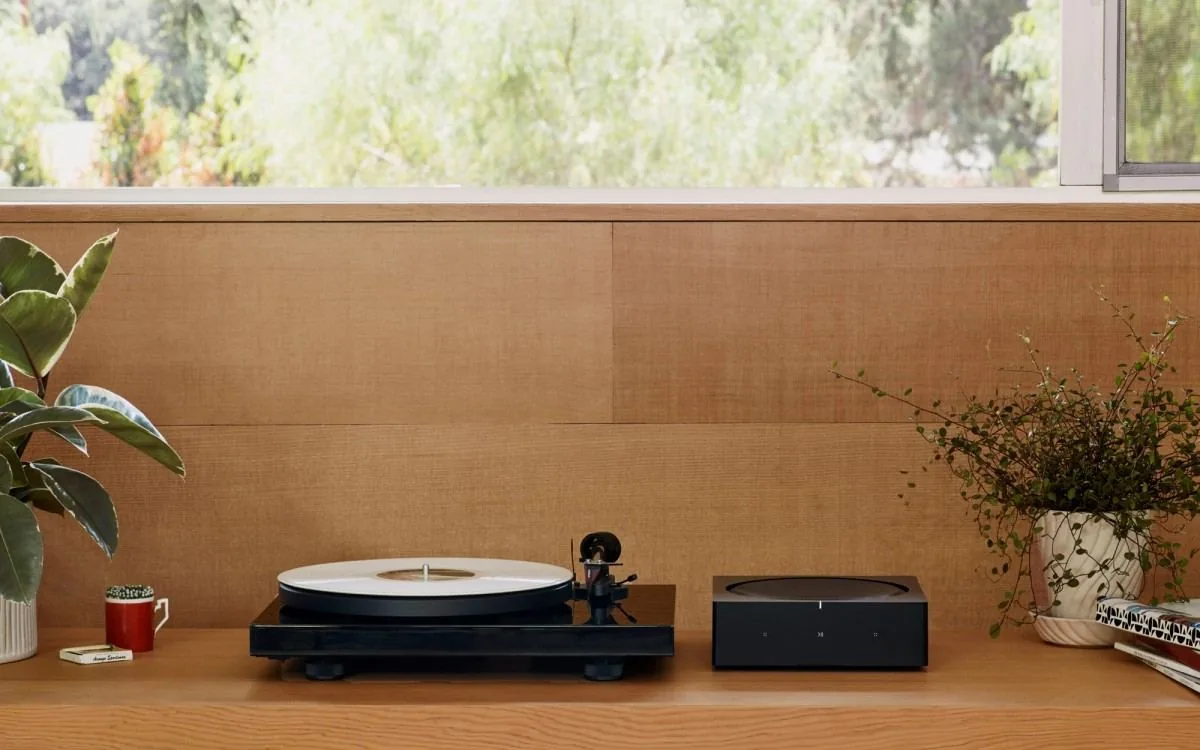 A turntable and a Sonos Amp on a wooden shelf by a window